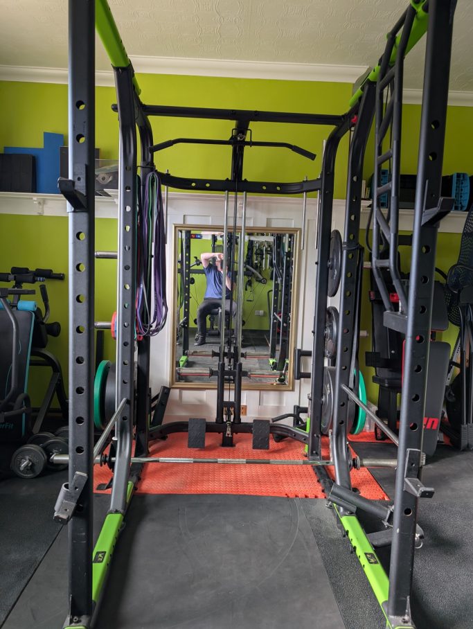 Home gym setup featuring a squat rack, weights, and a mirror. Green walls in the background.