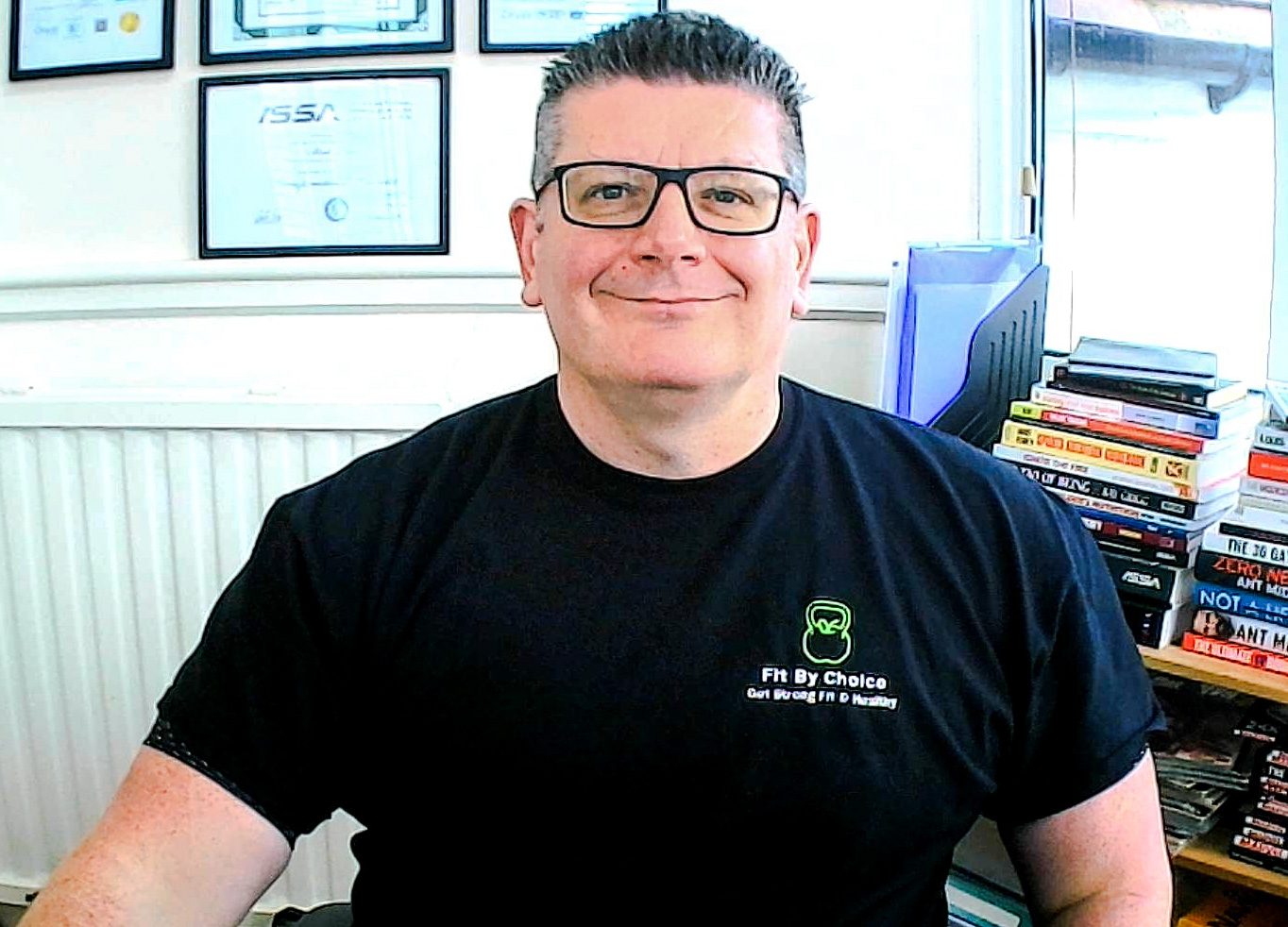 Mark Saker Coach Fit By Choice Smiling Mark Saker in a black t-shirt, seated in an office with books and certificates in the background.